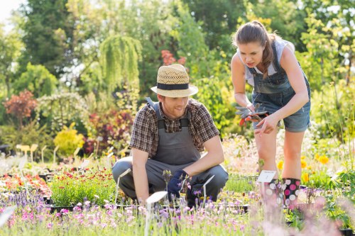 A gardener and a young girl are working together in a lush, well-maintained garden, surrounded by an array of colorful flowers, flowering shrubs, and tall trees. The gardener, dressed in a checked shirt, gardening apron, and a straw hat, is kneeling on freshly cut grass, carefully tending to the flower beds with gardening gloves on, while the girl, wearing a sleeveless top and denim shorts, is leaning over to assist with planting or weeding. The garden features neat flower borders with a variety of blooming perennial plants in shades of pink, purple, and yellow, set against a backdrop of vibrant green hedges, leafy bushes, and mature trees providing shade. The soil beds are well-weeded, and small garden tools and plant labels are visible nearby. Bright natural daylight illuminates the scene, suggesting a clear, sunny day, ideal for outdoor gardening activities. This scene reflects professional gardening services offered by Landscapers Feltham, emphasizing meticulous lawn, flower bed, and plant care within a landscaped outdoor space in Feltham or nearby TW postcode areas.