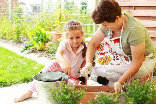 A woman and young girl are working together in a well-maintained garden, planting or tending to potted plants in a backyard outdoor space near a wooden shed. The woman, dressed in a light green top and a white apron with a colorful floral pattern, is wearing gardening gloves and appears to be assisting the girl, who is sitting on the grass and wearing casual clothes with pink and white striped leggings. They are using gardening tools, such as a small trowel, and handling soil or compost in rectangular plant containers, which are positioned on the patio area. The garden features lush green lawn, flowering plants, and a variety of shrubs and trees in the background, all bathed in natural light under a partly sunny sky. The scene reflects a caring, outdoor gardening activity typical of a residential garden maintained with care, aligning with professional gardening and landscaping services offered by companies like Landscapers Feltham, serving local communities in the Feltham area and TW postcode district.