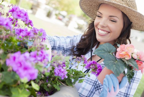 A woman wearing a straw hat and gardening gloves is smiling while tending to colorful flowering plants in a well-maintained garden. The garden features a variety of vibrant purple, pink, and orange blooms arranged in flower beds and containers, with lush green foliage providing a natural backdrop. The lawn area in the background is neatly trimmed, with a combination of grass and soil patches visible. Sunlight filters through the trees, casting a bright and cheerful atmosphere suitable for outdoor gardening activities. The scene captures a peaceful moment of garden care, reflecting professional landscaping and gardening services available in Feltham, with a focus on creating vibrant, healthy outdoor spaces.
