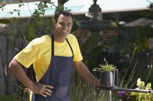 A man wearing a yellow T-shirt and a dark apron stands in a landscaped garden, smiling at the camera. He is holding a black plant pot containing a young, green shrub with fine foliage. The garden features a neatly maintained lawn with dense, green grass in the foreground. In the background, there are various plants and shrubs, some with yellow flowers, and a wooden fence partially covered with climbing plants. A garden ornament or fountain is visible behind the man, and there is a structure with a tiled roof, likely a garden shed or summerhouse, partially obscured by foliage. The scene is outdoors under natural daylight, with clear weather conditions, and suggests an environment suitable for gardening and outdoor landscape maintenance services, such as those offered by Landscapers Feltham in the Feltham area, TW13 postcode, emphasizing professional garden care and planting activities.