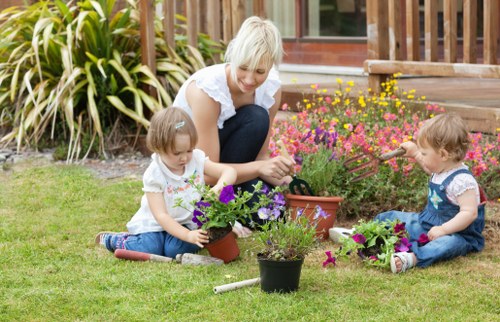 A garden scene in a residential outdoor space featuring a well-maintained grassy lawn with lush, dense green grass. In the foreground, a woman with short blonde hair wearing a white top and dark trousers is engaging with two young children, both with light brown hair and dressed in casual summer clothing, actively involved in planting and tending to small flowering plants in terracotta and plastic pots. The garden has vibrant flower beds with a variety of colourful blooms, including pink and purple flowers, situated near a wooden deck or steps leading to the house, which is partially visible in the background. Surrounding the garden are tall ornamental grasses and shrubs, adding natural texture to the scene. The weather appears bright and sunny, creating natural lighting that highlights the natural tones of the plants, soil, and garden features, reflecting the outdoor maintenance and landscaping services of Landscapers Feltham, with a focus on sustainable gardening practices.