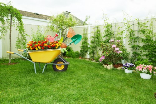 A well-maintained residential garden featuring a lush, green lawn with evenly cut grass occupying the foreground and extending towards a white wooden fence in the background. On the left side, there is a yellow wheelbarrow filled with various brightly coloured flowers, including red, yellow, and orange blooms. The garden is bordered by flower beds containing pink, purple, and white flowering plants, some in white pots, adding colour and texture to the space. Behind the flower beds, a row of leafy green shrubs and a small tree provide natural privacy and a sense of enclosure. The scene appears to be during daytime with soft natural lighting, and a partly cloudy sky overhead. The overall setting suggests a typical outdoor area that would benefit from professional gardening services such as lawn care, shrub trimming, and flower bed maintenance, aligning with the services offered by Landscapers Feltham. The environment is tidy, inviting, and shows signs of regular upkeep suitable for a residential outdoor space in Feltham, Middlesex.