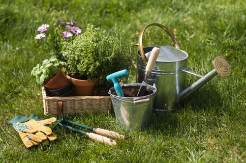 A neatly maintained backyard garden featuring a lush green lawn with dense, evenly cut grass. In the foreground, there are two small gardening tools, including a pair of pruning shears and a hand rake, placed on the grass. To the left, a woven basket holds a variety of potted plants and herbs, including flowering and leafy greenery, arranged in terracotta and plastic pots. Behind the tools and basket, a silver metal watering can with a curved handle and a long spout is positioned on the grass. The background includes a large shrub or hedge with dense foliage, contributing to a private and well-tended outdoor space. The scene suggests a pleasant, cloudy day, ideal for garden maintenance and planting activities, representative of professional gardening services available in Feltham, Middlesex, as promoted by Landscapers Feltham.
