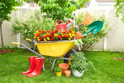 This outdoor garden scene features a green lawn with a central yellow wheelbarrow filled with bright red and yellow flowering plants, situated in front of a white fence. Behind the wheelbarrow, there are lush green shrubs and small trees providing a natural backdrop, with several gardening tools including a pink watering can and a blue and green spade resting among the plants. To the left, a pair of red rubber boots is placed on the grass, while in front of the wheelbarrow, there are small terracotta and plastic plant pots, some containing greenery. The garden appears well-maintained, with vibrant colors and a tidy arrangement, indicative of an outdoor space prepared for gardening or landscaping work, consistent with professional garden maintenance services offered by companies like landscapersfeltham.co.uk in the Feltham area. The scene is illuminated by natural daylight, suggesting mild weather conditions suitable for outdoor gardening activities.