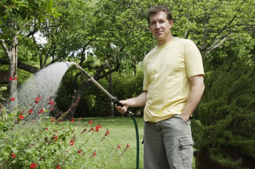A man with light brown hair wearing a pale yellow T-shirt and grey cargo trousers is standing in a well-maintained garden, holding a garden hose and watering plants. The garden features a lush green lawn in the foreground, edged by a border of blooming red flowers and leafy shrubs. Behind him, there are dense, mature trees with vibrant green foliage providing shade, as well as neatly trimmed hedges and bushes that create a structured landscape. The scene appears to be outdoors on a bright, slightly cloudy day, suggesting mild weather suitable for gardening. The garden layout includes a mix of grass, flowering plants, and timber fencing, with a background of tall trees and natural greenery. The image conveys a professional outdoor space that could benefit from lawn care and garden maintenance services offered by Landscapers Feltham, serving the local area near Feltham within the TW postcode district.
