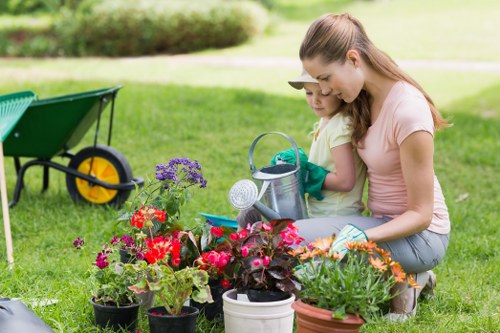 In a well-maintained garden, a woman and a young girl are engaged in gardening activities amidst an outdoor setting. The foreground features a variety of potted flowering plants with vibrant red, pink, and purple blossoms, placed on lush, green grass. To the left, a green wheelbarrow with yellow handles and a black wheel rests on the lawn, indicating recent or ongoing garden work. Behind the gardening duo, the background reveals a neatly trimmed hedge running horizontally across the scene, with a grassy lawn extending into the distance. The woman is kneeling on the grass, holding a watering can, while the girl, wearing gardening gloves, is attentively watering the plants, both having light skin and casual summer clothing. The environment appears bright and sunny, with natural daylight illuminating the scene, suggesting a pleasant weather day suitable for outdoor gardening. The overall setting exemplifies a well-kept, defining a peaceful outdoor space that could be part of a residential garden in Feltham, connecting to professional gardening services like those offered by Landscapers Feltham, especially focusing on garden planting and maintenance.