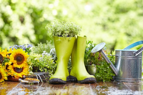 The image depicts a garden scene with a wooden outdoor table featuring a variety of gardening tools and decorative elements. In the center, there are a pair of lime green rubber gardening boots filled with white and green flowering plants, standing upright and serving as makeshift planters. To the left, a bunch of bright yellow sunflowers and other colorful flowers arrange a cheerful contrast against the natural wood surface. On the right side, a metallic watering can with a long spout is positioned, ready for watering plants, and nearby are small garden tools including a trowel and pruning shears. The background reveals a lush, green backyard with blurred foliage, suggesting a well-maintained outdoor space typical of a residential garden in Feltham, Middlesex. The scene is illuminated by natural daylight, enhancing the vibrant colours and textures of the plants and garden accessories, emphasizing a peaceful, neatly kept garden environment suitable for outdoor maintenance and landscaping activities by Landscapers Feltham.