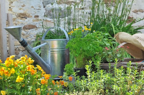 The image depicts a small, well-maintained garden area with a diverse collection of plants and garden elements. In the foreground, there are vibrant yellow and orange flowering plants, possibly marigolds or similar garden flowers, growing densely along the edge of the garden bed. Behind these, a patch of lush green foliage with small leaves adds complexity and fullness to the planting arrangement. Situated among the plants is a traditional metal watering can with a curved handle and a long spout, indicating ongoing garden care. To the right, a straw sun hat rests on the edge of the garden, suggesting the presence of a gardener or outdoor routine. The garden features a backdrop of a weathered stone wall, adding texture and a natural rustic charm to the scene. Tall green grass or reeds grow along the boundary, providing height and vertical interest. The scene is lit by natural daylight, with clear weather conditions, making the natural tones of the plants, soil, and garden accessories visible and vibrant. This outdoor space exemplifies typical landscaping and gardening elements that would be maintained by professional landscapers such as Landscapers Feltham, enhancing its aesthetic appeal and functionality within a small backyard or front garden setup near Feltham, Middlesex.