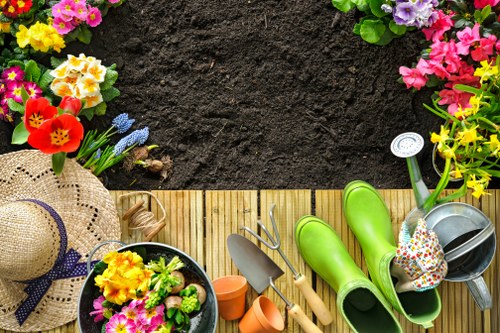 A recently prepared garden space with a focus on gardening tools and plants. The foreground features a wooden garden border, with a lush green lawn beyond. On the wooden surface, there are bright green rubber gardening gloves, a metal watering can, and a floral-patterned gardening hat with a wide brim. Nearby, a small black pot contains colourful flowering plants, including yellow and pink blossoms. Several empty terracotta pots are scattered around, hinting at ongoing planting activities. In the background, a patch of dark, well-prepared soil is bordered by vibrant flower beds filled with various flowering plants in shades of pink, purple, and yellow. The overall scene suggests a well-maintained outdoor space suitable for landscaping and gardening services in Feltham, with natural sunlight illuminating the garden during a clear day. The environment appears tidy and ready for planting or lawn care work, emphasizing a neat and inviting outdoor area typical of residential gardens in the region.