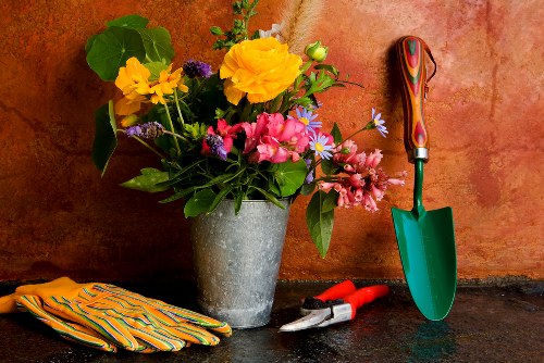 A close-up image of a gardening scene featuring a large, grey concrete flowerpot filled with a variety of colorful flowering plants, including yellow, pink, and purple blooms, set against a warm, textured orange wall. In front of the pot, on a dark stone surface, there are a pair of gardening shears and a small trowel with a green handle, along with a pair of striped gardening gloves. The scene suggests outdoor gardening activity, with a focus on planting and flower care, reflecting typical garden maintenance tasks in a residential outdoor space near Feltham, with possible relevance to local garden makeover services provided by Landscapers Feltham.