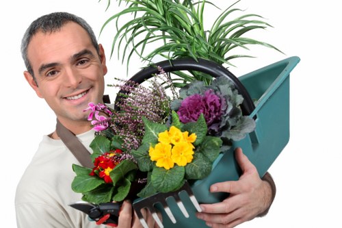 A man with short dark hair and a friendly expression is holding a green plastic garden basket filled with a variety of potted plants and flowers, including purple, yellow, and red blooms. The basket contains a potted fern with long, arching green fronds and a small, lush shrub. The background is plain and light-colored, emphasizing the garden elements and the man's attire, which is casual. The scene suggests outdoor or garden maintenance service in a typical residential garden setting, with natural natural lighting. The attention to detail in the arrangement reflects careful gardening and plant care, suitable for visual descriptions on a landscaping or gardening service website servicing Feltham and the surrounding areas in West London. This image aligns with landscaping themes, showcasing plant handling and garden maintenance.