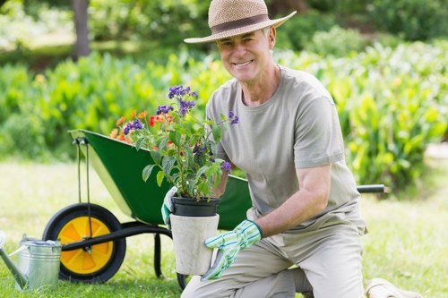 A smiling man wearing a wide-brimmed straw hat, grey T-shirt, and gardening gloves is kneeling on a neatly maintained lawn in a garden. He is holding a container with purple and red flowering plants, preparing to plant them into the garden. Behind him, there is a green wheelbarrow with large yellow and black wheels, and a metal watering can placed on the grass nearby. The background features a lush, green hedge, with tall trees providing natural shade, and the garden is bathed in soft natural daylight indicating clear weather. The scene depicts outdoor gardening activity within a well-kept landscaped yard, emphasizing lawn care, planting, and general garden maintenance typical of a professional landscaping service in Feltham, close to the TW13 postcode area.