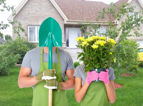 A garden scene in a residential area with a well-maintained front lawn and a backdrop of a brick house with white window frames and a gabled roof. In the foreground, two gardeners are visible; one wearing a bright green gardening apron and gloves, holding a silver spade with a teal plastic cover over the handle, while the other, also dressed in a grey shirt and pink gloves, is holding a large pink pot filled with vibrant yellow flowering plants. The garden features neatly cut grass, small shrubs, and delineated flower beds with soil visible along the edges, suggesting recent planting activity. The environment appears bright and sunny, indicating clear weather, ideal for outdoor gardening work. The scene reflects professional gardening and planting services, highlighting the process of preparing the outdoor space, with an emphasis on creating a lush, inviting garden landscape typical of Feltham or surrounding areas in West London, serviced by Landscapers Feltham.