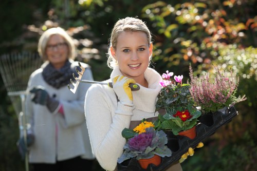 A young woman with blond hair tied back, smiling at the camera while holding a black plastic tray filled with flowering plants and garden tools, standing in a well-maintained backyard garden during daylight. Behind her, an older woman with blond hair and glasses, also smiling, is slightly blurred and appears to be engaging in gardening activities, such as pruning or planting. The garden features a lush green lawn with dense foliage, including shrubs and trees with green and reddish leaves, and a background of sunlight filtering through the foliage, creating a warm and inviting outdoor environment ideal for gardening and landscaping services in Feltham. The scene captures a bright, clear day with natural lighting, emphasizing the vibrant colours of the plants and the healthy appearance of the lawn, reflecting the nurturing nature of professional gardening work by Landscapers Feltham.