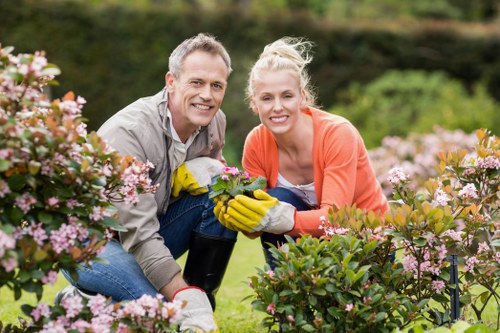 A man and a woman are kneeling together in a lush garden, surrounded by flowering plants and shrubs with pink and white blooms. They are both smiling and wearing gardening gloves, with the woman holding a small potted plant. The garden features a well-maintained lawn with vibrant green grass, bordered by a variety of garden beds and leafy bushes, set against a background of trees and a hedge. The scene is outdoors on a bright, clear day, suggesting a pleasant weather condition ideal for gardening activities. This image reflects a landscaped garden environment typical of residential outdoor spaces in Feltham, showcasing elements like flowering bushes, trimmed hedges, and a tidy lawn that align with professional gardening and landscaping services offered by Landscapers Feltham in the local area. It highlights the joy of gardening and outdoor maintenance in a natural setting, emphasizing the importance of garden care and plant nurturing within a thoughtfully designed garden landscape.